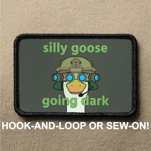 May include: A black and green patch with a cartoon goose wearing a helmet and night vision goggles. The text "silly goose going dark" is printed on the patch.