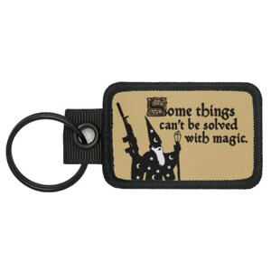 Könnte beinhalten: Ein schwarzer und brauner Schlüsselanhänger mit einer Schwarzweißillustration eines Zauberers, der eine Waffe und einen Zauberstab hält. Der Text auf dem Schlüsselanhänger lautet "Some things can't be solved with magic."