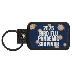 Può includere: Un portachiavi nero con un'etichetta rettangolare blu e bianca che dice "2025 Bird Flu Pandemic Survivor". L'etichetta è decorata con un motivo di virus stilizzati rosa e gialli.