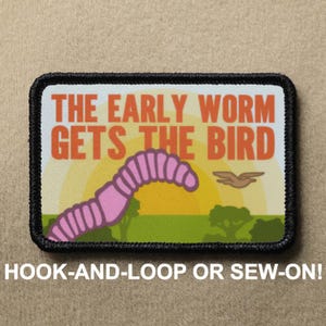May include: A pink worm with a black outline is arched over a yellow sun with a brown bird flying above it. The text "THE EARLY WORM GETS THE BIRD" is in orange letters on a white background. The patch is black with white stitching around the edges. The text "HOOK-AND-LOOP OR SEW-ON!" is below the patch.
