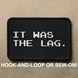 Puede incluir: Parche negro con texto blanco que dice "IT WAS THE LAG." El parche tiene un borde negro y está diseñado para ser fijado con velcro o cosido.