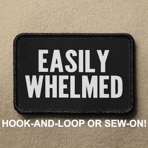 May include: Black patch with white text that reads "EASILY WHELMED". The patch has a black border and is designed to be attached with hook-and-loop or sewn on.