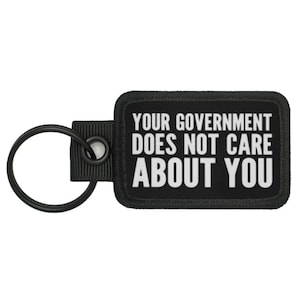 Könnte beinhalten: Schwarzer Schlüsselanhänger mit weißem Text, der "Ihre Regierung kümmert sich nicht um Sie" lautet.