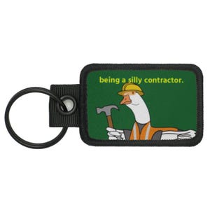 Puede incluir: Un llavero negro y verde con un ganso blanco que lleva un casco amarillo y un chaleco naranja. El ganso sostiene un martillo y el texto "being a silly contractor." está impreso en el llavero.