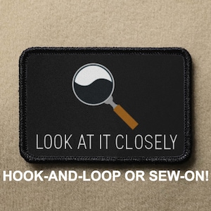 May include: Black patch with a white magnifying glass with a brown handle. The text "LOOK AT IT CLOSELY" is printed in white below the magnifying glass.