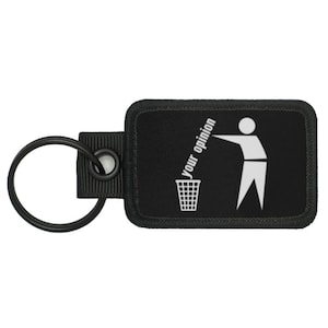 May include: Black rectangular keychain with a metal ring. The keychain features a white graphic of a person throwing something into a trash can, with the words "your opinion" in white.
