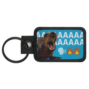 May include: A rectangular keychain with a black border features a brown sea lion with an open mouth on a blue background. The text "AAAAA" is above "AAAAA". A white head with sound waves and three flame emojis are also present.