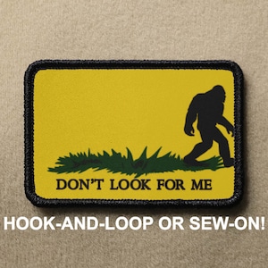 Puede incluir: Parche amarillo con una silueta negra de Bigfoot caminando lejos de un parche de hierba verde. El texto "DON'T LOOK FOR ME" está impreso en negro debajo de la imagen.