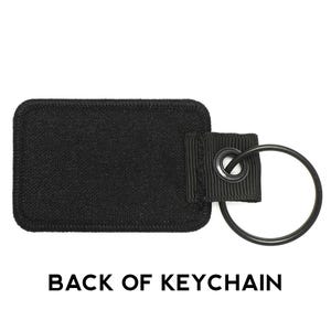 May include: Black fabric keychain with a black metal ring. The back of the keychain is blank.