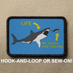 Op de afbeelding: Een blauwe en witte patch met een cartoonhaai die een kleinere vis achtervolgt. De tekst "LIFE" staat boven de haai met een pijl die ernaar wijst. De tekst "MY HOPES AND DREAMS" staat onder de vis met een pijl die ernaar wijst. De patch is zwart met een witte rand en zegt "HOOK-AND-LOOP OR SEW-ON!" onderaan.