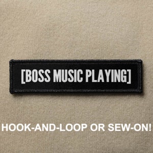 Pode incluir: Remendo preto com texto branco que diz "[BOSS MUSIC PLAYING]" e as palavras "HOOK-AND-LOOP OR SEW-ON!" abaixo.