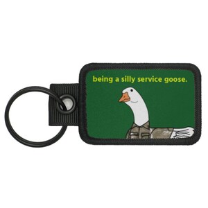 May include: A black key chain with a green patch featuring a white goose wearing a camouflage uniform. The text on the patch reads "being a silly service goose."