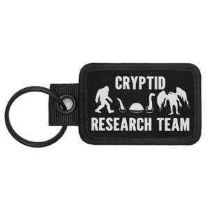 May include: Black key chain with a black patch that says "Cryptid Research Team" with a white silhouette of a Bigfoot, a Loch Ness Monster, and a Mothman.