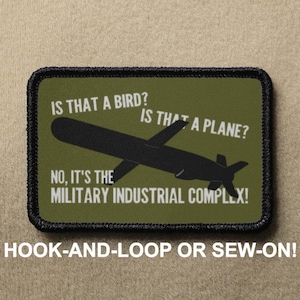 May include: A green military-style patch with black stitching. The patch features a black silhouette of a missile with the text "Is that a bird? Is that a plane? No, it's the Military Industrial Complex!" in white.