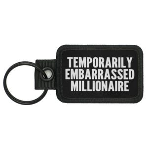 Puede incluir: Llavero negro con una etiqueta rectangular negra que dice "Temporarily Embarrassed Millionaire" en letras blancas.