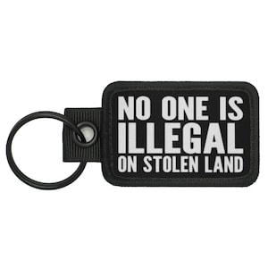 Pode incluir: Chaveiro preto com uma etiqueta retangular preta que diz "NO ONE IS ILLEGAL ON STOLEN LAND" em letras brancas.