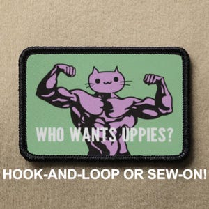 Peut inclure: Un écusson rectangulaire à bordure noire présente un personnage de chat musclé en violet fléchissant ses bras sur un fond vert clair. Le texte "WHO WANTS UPPIES?" est en dessous du chat. Le texte "HOOK-AND-LOOP OR SEW-ON!" est en bas.