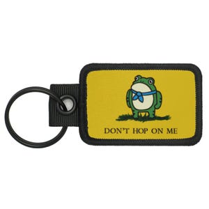 May include: A rectangular keychain with a yellow background and a cartoon frog wearing a blue neckerchief. The text "DON'T HOP ON ME" is below the frog. The keychain has a black border and a black key ring.