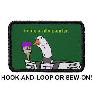 May include: A black and white goose with purple spots is holding a paintbrush with purple paint. The goose is wearing a black and white uniform. The text "being a silly painter." is above the goose. The image is on a green background and is surrounded by a black border. The text "HOOK-AND-LOOP OR SEW-ON!" is below the image.