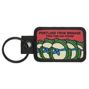 May include: A rectangular keychain patch with a black border and a red background. The patch features a cartoon illustration of five green frogs with blue bows. The text "PORTLAND FROG BRIGADE" and "FROG TOGETHER STRONG" are printed on the patch. A black key ring is attached.
