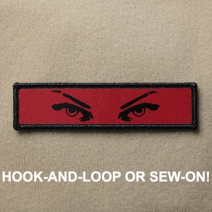 Puede incluir: Parche rectangular rojo con ojos bordados en negro. El parche tiene costuras negras alrededor de los bordes. El texto "HOOK-AND-LOOP OR SEW-ON!" está impreso debajo del parche.