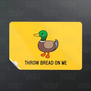 Op de afbeelding: Gele sticker met een cartoon eend en de tekst "THROW BREAD ON ME".