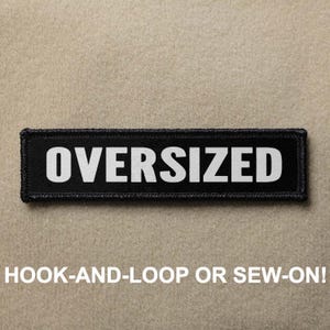 Puede incluir: Parche negro con texto blanco que dice "OVERSIZED". El parche está diseñado para ser fijado con velcro o cosido.