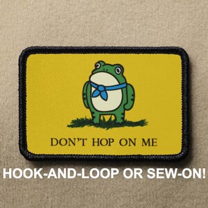 Puede incluir: Parche rectangular con fondo amarillo y una rana verde con pañuelo azul. El texto "DON'T HOP ON ME" está debajo de la rana. El parche tiene un borde negro y el texto "HOOK-AND-LOOP OR SEW-ON!" en la parte inferior.