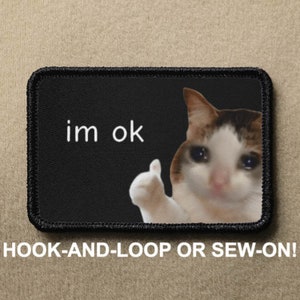 May include: A black and white patch with a cat giving a thumbs up and a sad expression. The text "im ok" is printed on the patch. The patch is for hook-and-loop or sew-on.