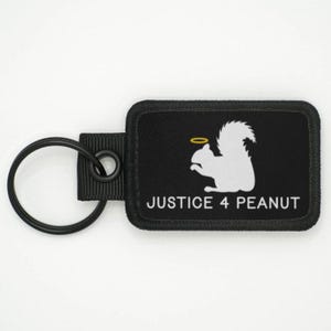 Può includere: Portachiavi rettangolare nero con uno scoiattolo bianco con un'aureola e la scritta "JUSTICE 4 PEANUT". Il portachiavi ha un anello di metallo nero e un passante in tessuto nero.