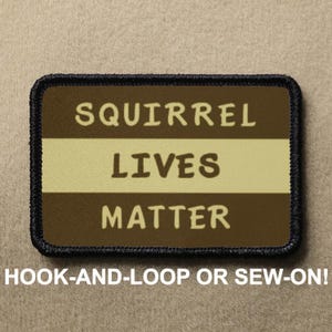 May include: A brown and tan patch with black stitching around the edges. The patch has the text "Squirrel Lives Matter" in white lettering.