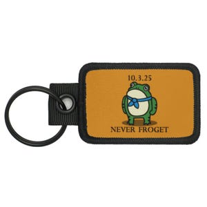 May include: A rectangular keychain with a black border and a cartoon frog design. The frog is green with a blue neckerchief, standing on an orange background. The text "10.3.25" and "NEVER FROGET" are above and below the frog.