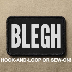 Pode incluir: Patch preto com texto branco que diz "BLEGH". O patch é para fixação de velcro ou para costurar.