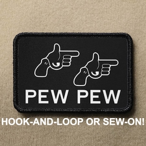 Puede incluir: Parche negro con contornos blancos de dos manos apuntando y el texto "PEW PEW". El parche es para fijación de gancho y bucle o para coser.