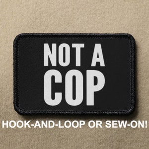 May include: Black patch with white text that reads "NOT A COP". The patch is for hook-and-loop or sew-on attachment.