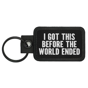 以下が含まれることがあります： 黒いキーホルダーに、黒い布製のタグが付いており、白い文字で「I got this before the world ended」と書かれています。