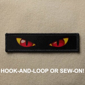 Può includere: Toppa nera con due occhi rossi con pupille gialle. La toppa ha cuciture nere attorno ai bordi. Il testo "HOOK-AND-LOOP OR SEW-ON!" è sotto la toppa.