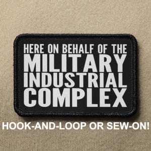 Puede incluir: Parche negro con texto blanco que dice "HERE ON BEHALF OF THE MILITARY INDUSTRIAL COMPLEX". El parche tiene un borde negro y está diseñado para ser fijado con velcro o cosido.