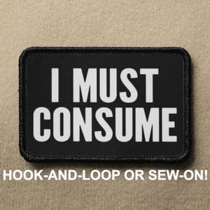 Peut inclure: Patch noir avec du texte blanc qui dit "I MUST CONSUME". Le patch est pour une fixation à boucle ou à coudre.