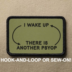 May include: A green and black patch with a white outline. The patch features a circular arrow with the text "I WAKE UP" and "THERE IS ANOTHER PSYOP". The patch is labelled "HOOK-AND-LOOP OR SEW-ON!"