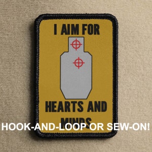 Può includere: Una toppa gialla con cuciture nere attorno ai bordi. La toppa presenta un bersaglio con due centri rossi e il testo "I AIM FOR HEARTS AND MINDS" in nero. Il testo "HOOK-AND-LOOP OR SEW-ON!" è stampato sotto il bersaglio.