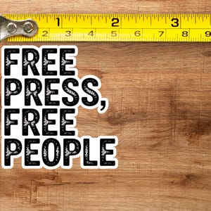 May include: A white sticker with the text "FREE PRESS, FREE PEOPLE" in a bold, black font. The sticker is placed on a wooden surface, with a yellow measuring tape in the frame. The tape measure shows inches, equivalent to cm.