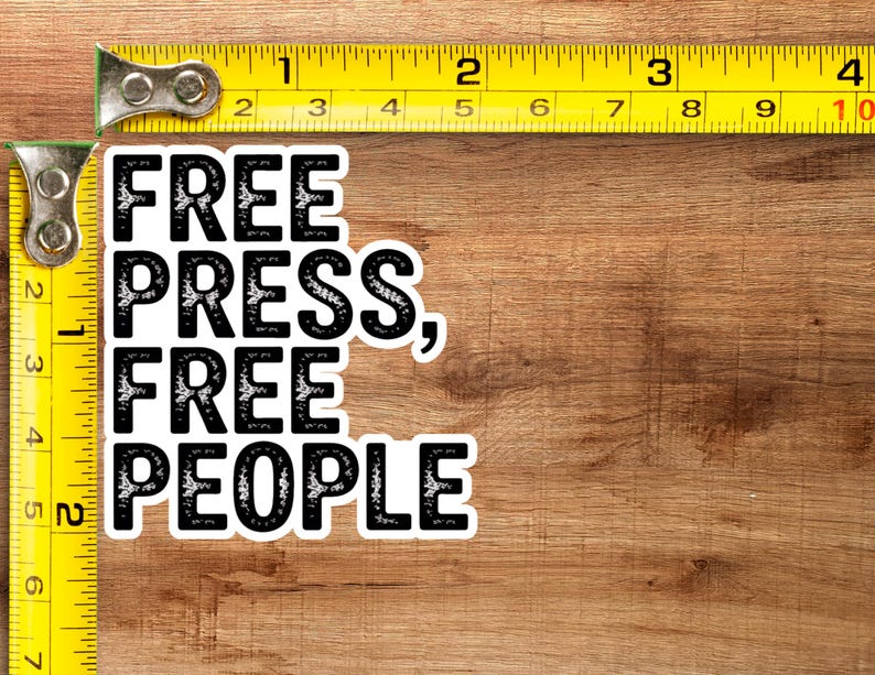 May include: A white sticker with the text "FREE PRESS, FREE PEOPLE" in a bold, black font. The sticker is placed on a wooden surface, with a yellow measuring tape in the frame. The tape measure shows inches, equivalent to cm.
