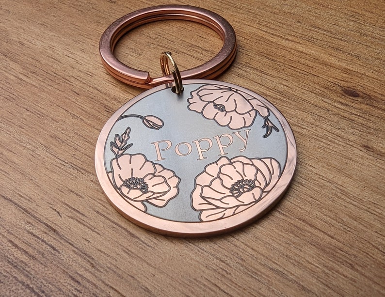 Pu&ograve; includere: Un medaglietta rotonda per animali domestici, color rame, con il nome "Poppy" inciso al centro. La medaglietta presenta un design floreale con fiori di papavero sul bordo e un portachiavi abbinato.
