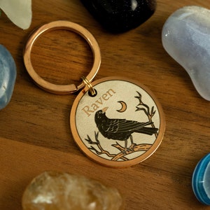 Op de afbeelding: Een koperen sleutelhanger met een rond label waarop "Raven" staat en een zwarte raaf met een halve maan op de achtergrond.