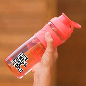 May include: A clear, pink water bottle with a matching pink lid and handle. The bottle has the words "FREE PRESS, FREE PEOPLE" in a black, stacked font. The bottle is held by a person.
