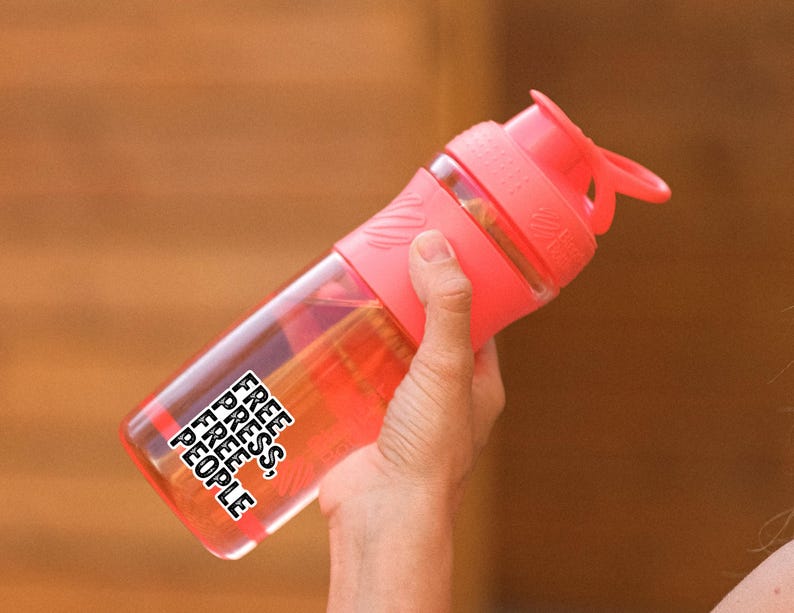 May include: A clear, pink water bottle with a matching pink lid and handle. The bottle has the words "FREE PRESS, FREE PEOPLE" in a black, stacked font. The bottle is held by a person.
