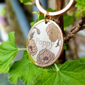 Pu&ograve; includere: Un medaglietta rotonda dorata per animali domestici con il nome "Poppy" inciso al centro. La medaglietta presenta un design floreale con tre fiori di papavero di colore marrone chiaro. La medaglietta &egrave; attaccata a un anello dorato.