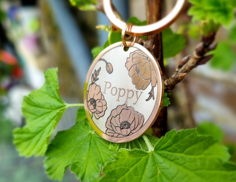 Pu&ograve; includere: Un medaglietta rotonda dorata per animali domestici con il nome "Poppy" inciso al centro. La medaglietta presenta un design floreale con tre fiori di papavero di colore marrone chiaro. La medaglietta &egrave; attaccata a un anello dorato.
