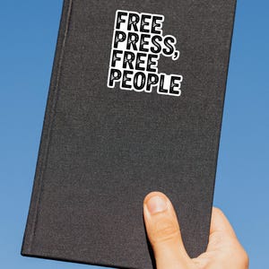 May include: A black book with the words "FREE PRESS, FREE PEOPLE" in a white, bold font. The book is held by a hand against a clear blue sky.